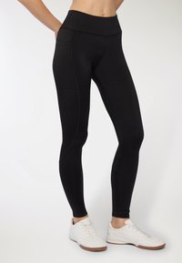 Czarne legginsy o wysokim stanie z gładką teksturą, z kieszeniami po bokach i kontrastowym przeszyciem, w połączeniu z białymi butami sportowymi.