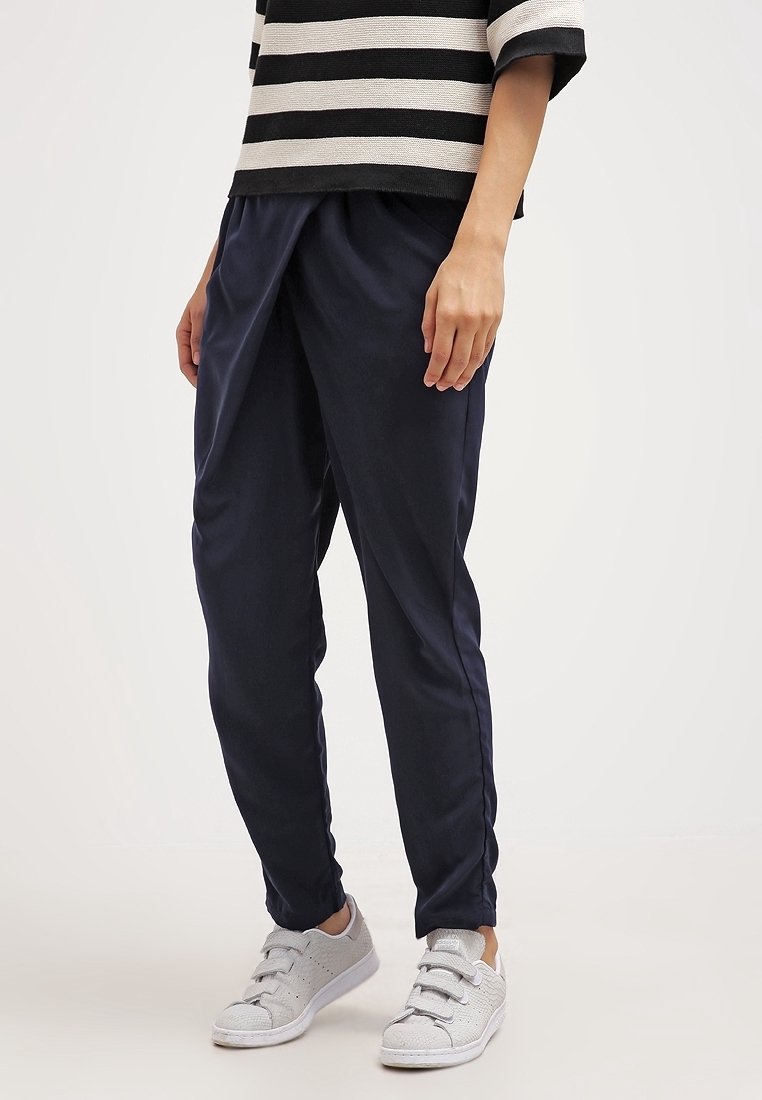 Personne portant un pantalon bleu marine fuselé, un haut rayé noir et blanc, et des baskets blanches, debout devant un fond uni.