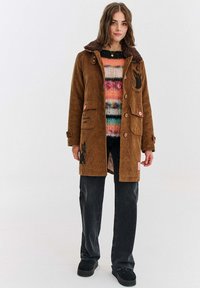 Brązowy sztruksowy płaszcz z kołnierzem z futra syntetycznego, dwoma przednimi kieszeniami, różowymi guzikami i czarnymi motywami zwierzęcymi. Noszony na wielokolorowym swetrze robionym na drutach.