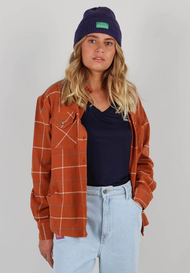 Chemise en flanelle rouille avec un motif à carreaux blancs, deux poches poitrine ; portée par-dessus un t-shirt en V bleu marine et associée à un jean bleu clair.