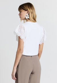 Top bianco a maniche corte con maniche in pizzo intricato, abbinato a pantaloni marrone chiaro. Tessuto morbido e vestibilità casual.