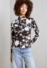 Langermet topp med et svart og hvitt blomstermønster, laget av stretchmateriale med høy hals og utskjæring. Kombinert med lyseblå jeans.