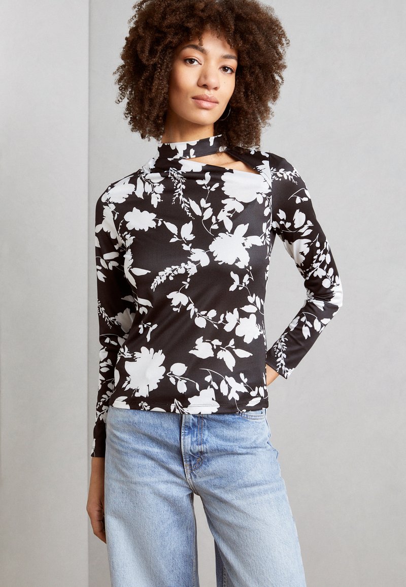 Langermet topp med et svart og hvitt blomstermønster, laget av stretchmateriale med høy hals og utskjæring. Kombinert med lyseblå jeans.