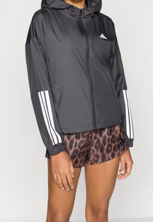Personne portant une veste à capuche noire zippée avec des bandes blanches sur les manches et un short marron à imprimé léopard avec un logo noir.