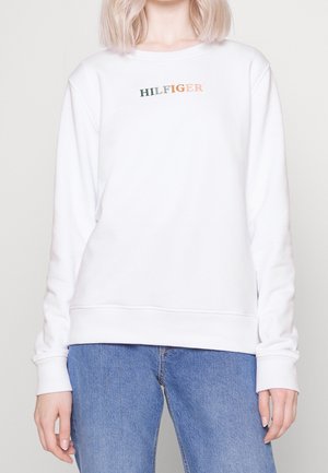 Il maglione bianco a collo rotondo presenta il logo ricamato "HILFIGER" in verde e arancione, con tessuto morbido e polsini e orlo a costine. Abbinato a un paio di jeans blu.