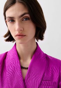 Chaqueta rosa con un patrón de cebra texturizado, que presenta un cuello con muesca y un bolsillo. El modelo lleva un collar de cuentas negras.