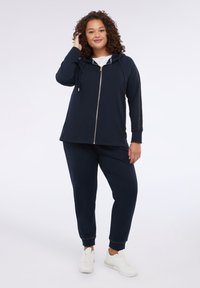 Granatowy hoodie na zamek z troczkami, w połączeniu z dopasowanymi spodniami. Miękka tkanina, ściągacze na rękawach i białe trampki dopełniają swobodny wygląd.