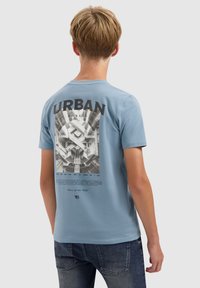 Jong persoon met kort blond haar die een lichtblauwe T-shirt draagt met een zwart-wit print van stedelijke architectuur en de tekst "URBAN JUNGLE" op de achterkant.