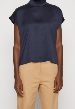 Persona con camiseta azul marino sin mangas con cuello alto y pantalones beige, de pie frente a un fondo claro y liso.