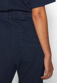 Mörkblå jumpsuit med korta ärmar, synliga sömmar och en bakficka med lock och knappstängning. Slät textur.