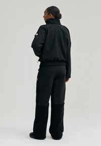 Fekete dzseki bolyhos gallérral és mandzsettával, fekete gyapjúbéléses nadrággal párosítva, mindkettő laza fazonnal és minimális díszítéssel.