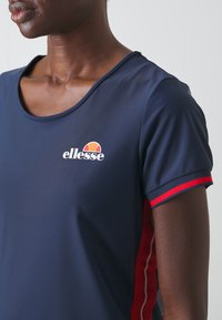 Camisa atlética de color navy con mangas cortas, que presenta un acento de rayas rojas y el logotipo "ellesse" en el pecho. Textura de tejido sintético suave.