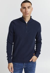 Tamsiai mėlynas zip-neck megztinis su raukšlėta tekstūra, su auksiniu užtrauktuku krūtinės srityje ir logotipo etikete kairiajame apačiame kampe.