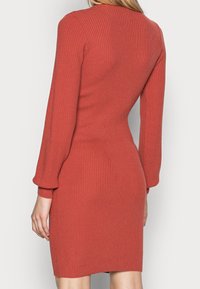 Abito a maglia a coste in arancione bruciato. Presenta maniche lunghe con polsini aderenti e una silhouette aderente, che termina sopra il ginocchio.