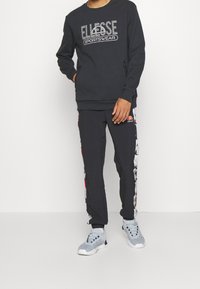 Sweat-shirt gris foncé avec le logo "Ellesse Sportswear", associé à un pantalon noir avec des panneaux latéraux colorés et des baskets grises.