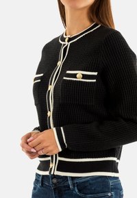 Cardigan nero in maglia con superficie testurizzata, dettagli bianchi lungo i bordi, bottoni dorati e due tasche frontali.