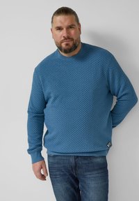 Sweter o fakturze w kolorze niebieskim z okrągłym dekoltem i prążkowanymi mankietami, zestawiony z ciemnymi dżinsami. Posiada charakterystyczny wzór i metkę z logo na dole.