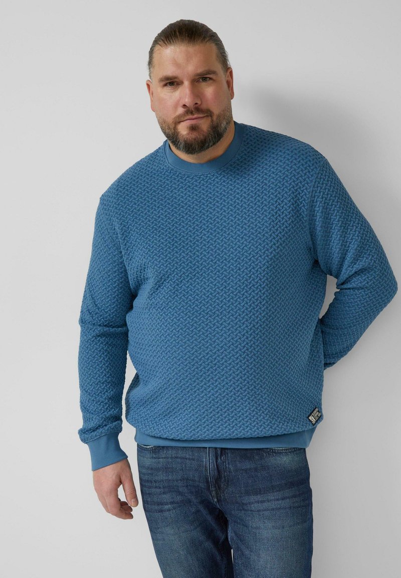 Sweter o fakturze w kolorze niebieskim z okrągłym dekoltem i prążkowanymi mankietami, zestawiony z ciemnymi dżinsami. Posiada charakterystyczny wzór i metkę z logo na dole.