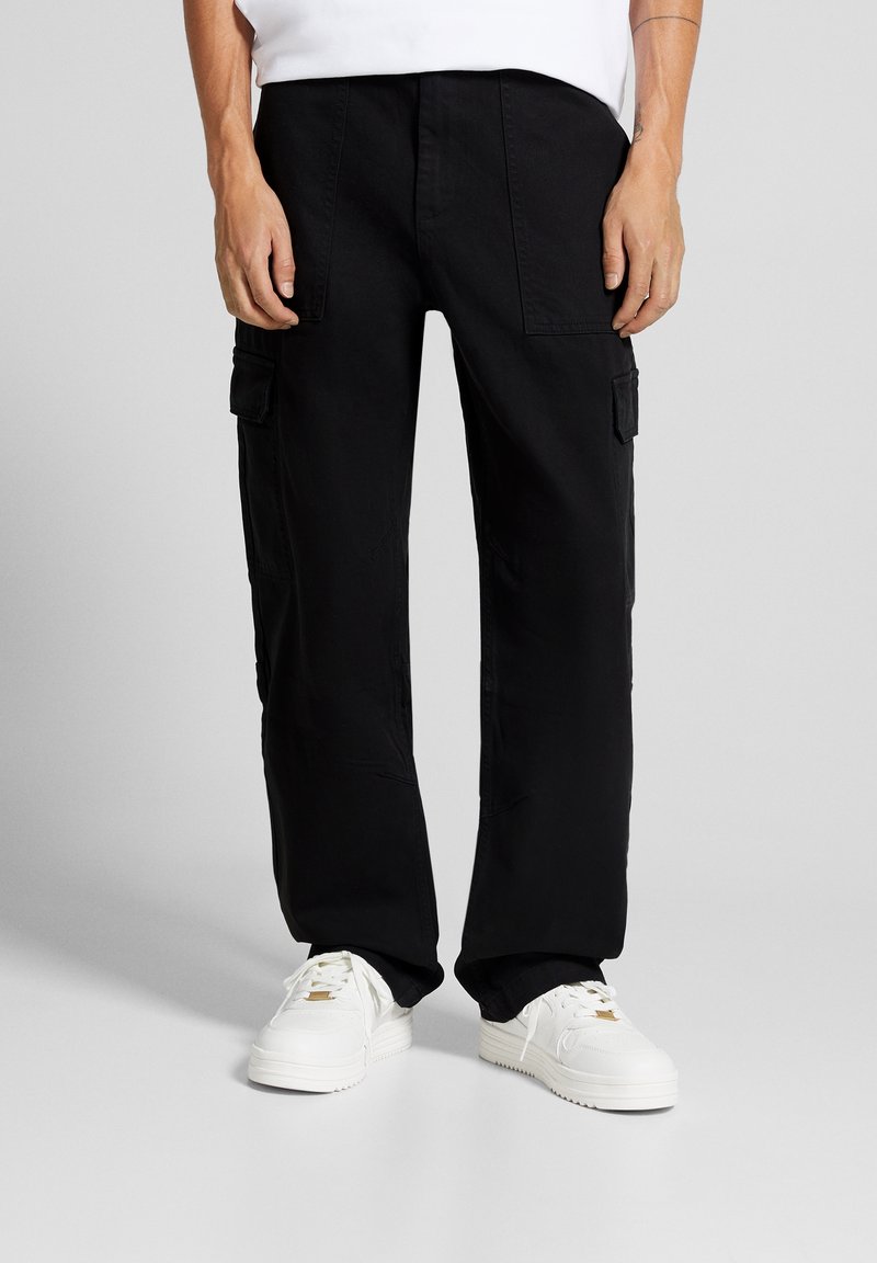 Homme portant un pantalon cargo noir et des baskets blanches debout sur un sol et un fond gris clair unis.