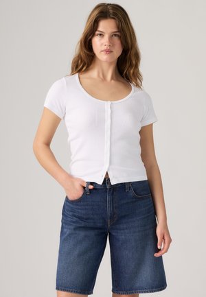 Jeune femme aux cheveux bruns ondulés portant un haut blanc boutonné à manches courtes et un short en jean bleu foncé au niveau des genoux sur un fond uni.