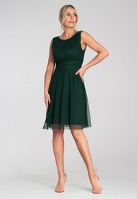 Grünes ärmelloses Kleid mit einem taillierten Oberteil, gefälteltem Detail und einem halbtransparenten Tüllrock. Entworfen mit einer klassischen Silhouette und subtiler Glanz.