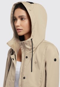 Beige hooded jacket with black drawstrings, featuring snap buttons and a logo on the sleeve. Smooth texture and fitted design.