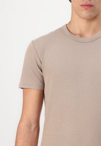 Beige t-paita lyhyillä hihoilla, joka on valmistettu ribkudotusta kankaasta; pyöreä kaula-aukko, vartaloa myötäilevä leikkaus, sileä rakenne ja siistit saumat ilman näkyviä logoja.