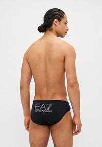 Schwarze Schwimmbriefs mit dem EA7 Emporio Armani Logo in einem Verlaufdesign auf der Rückseite. Glatte Textur, figurbetontes Design.