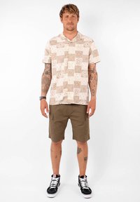 Chemise à manches courtes avec un motif de patchwork beige, portée avec un short cargo olive, associée à des baskets montantes noir et blanc.