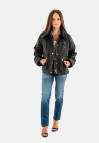 Zwarte waterdichte jas met een kraaghalslijn, gouden hardware en twee grote voorzakken. Draagt over een witte top en blauwe jeans.
