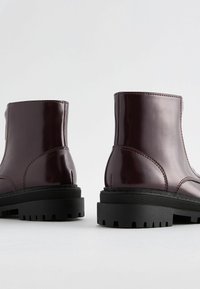 Bottines à patin bordeaux en vernis avec une semelle en caoutchouc noir épaisse, présentant une finition lisse et des coutures visibles le long des côtés.