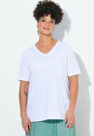 T-shirt en coton blanc avec un col en V, des manches courtes et des nœuds en crème décoratifs. Associé à une jupe plissée verte.