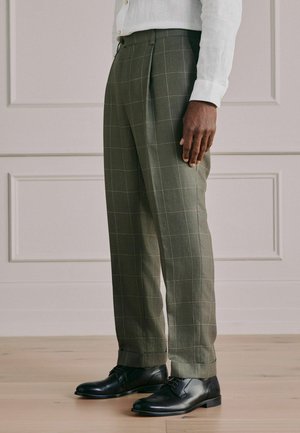 Man wearing olive-green checked dress pants, white shirt, and black leather dress shoes, standing on a wooden floor.