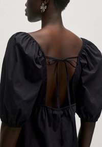 Schwarzes Kleid mit puffvollen Ärmeln, einem tiefen, offenen Rückenteil und einem gebundenen Detail. Der Stoff scheint leicht und mit einer glatten Textur zu sein.