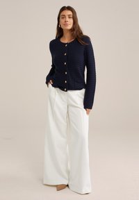 Navy ribbed cardigan with golden buttons and pockets, paired with white wide-leg trousers and nude heels, showcasing textured fabric.