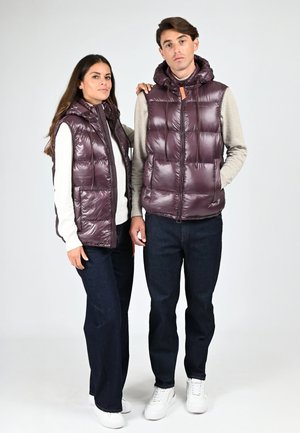 Un homme et une femme se tenant côte à côte, portant des gilets matelassés brillants violets assortis, des tops à manches longues, des jeans foncés et des baskets blanches.