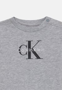 Grå bomull t-skjorte med rund hals, med et stort svart "CK"-logo og "Calvin Klein"-tekst i hvitt under logoen. Trykknappdetalj.