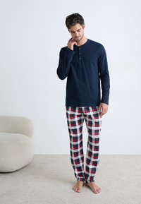 Man in een marineblauwe henley shirt en rode, witte en groene geruite pyjamabroek die blootsvoets op een beige tapijt staat naast een crèmekleurige ronde stoel.