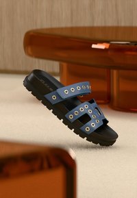 Μπλε λαστιχένιο σανδάλι με δύο φαρδιές λουρίδες που έχουν κυκλικά μεταλλικά περφορέ, υφασμένο μαύρο πάτο και σχέδιο με ανοιχτά δάχτυλα.