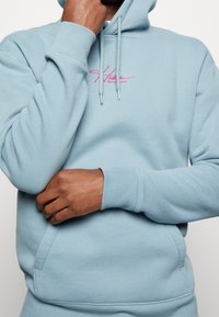 Sudadera azul claro de tela suave, con un bolsillo canguro frontal, capucha con cordones y un logo bordado en color rosa que contrasta.
