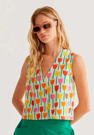 Mujer con cabello rubio ondulado lleva gafas de sol beige de gran tamaño, top sin mangas con escote en V y estampado de tulipanes coloridos, y pantalones verdes de cintura alta.