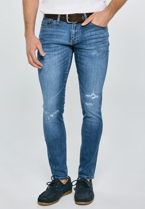 Mees, kes kannab kitsaid siniseid teksaseid kulunud detailidega, pruuni vööd, valget särki ja tumelillasid paeltega kingi, seisab ühe käega taskus.