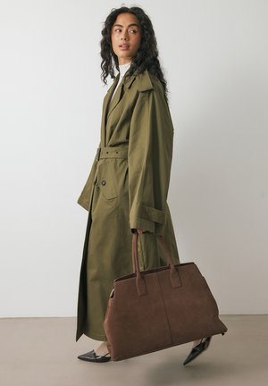Femme dans un trench-coat ceinturé olive, portant un grand sac fourre-tout marron, chaussée de chaussures noires à bout pointu, se tenant devant un fond clair et uni.