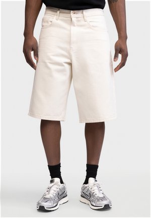 Άνδρας που φοράει off-white σορτς μέχρι τα γόνατα, μαύρες κάλτσες και ασημί-λευκά αθλητικά παπούτσια Nike, στέκεται μπροστά σε απλό φόντο.