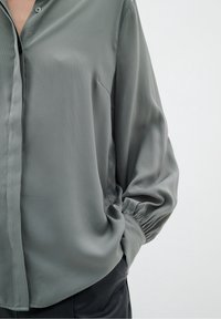 Blusa grigia chiara con una texture liscia, chiusura frontale con bottoni e polsini arricciati. Vestibilità sartoriale con delicate pieghe.
