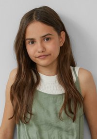 Kind mit langen, welligen braunen Haaren, das ein grünes ärmelloses Kleid über einem weißen Strickoberteil trägt. Hintergrund ist eine hellgraue Wand.