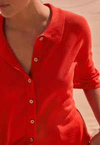 Roter Strickcardigan mit Kragen, kurzen Ärmeln und sieben Knöpfen mit floralen Akzenten. Weiche Textur und figurbetontes Design.