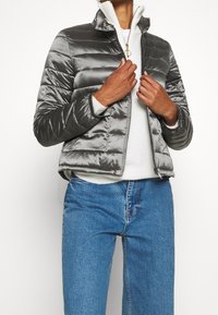 Lysegrå dunjakke med skinnende finish, vandret syning, stående krave og lynlåslukning. Båret over en hvid sweater og blå jeans.