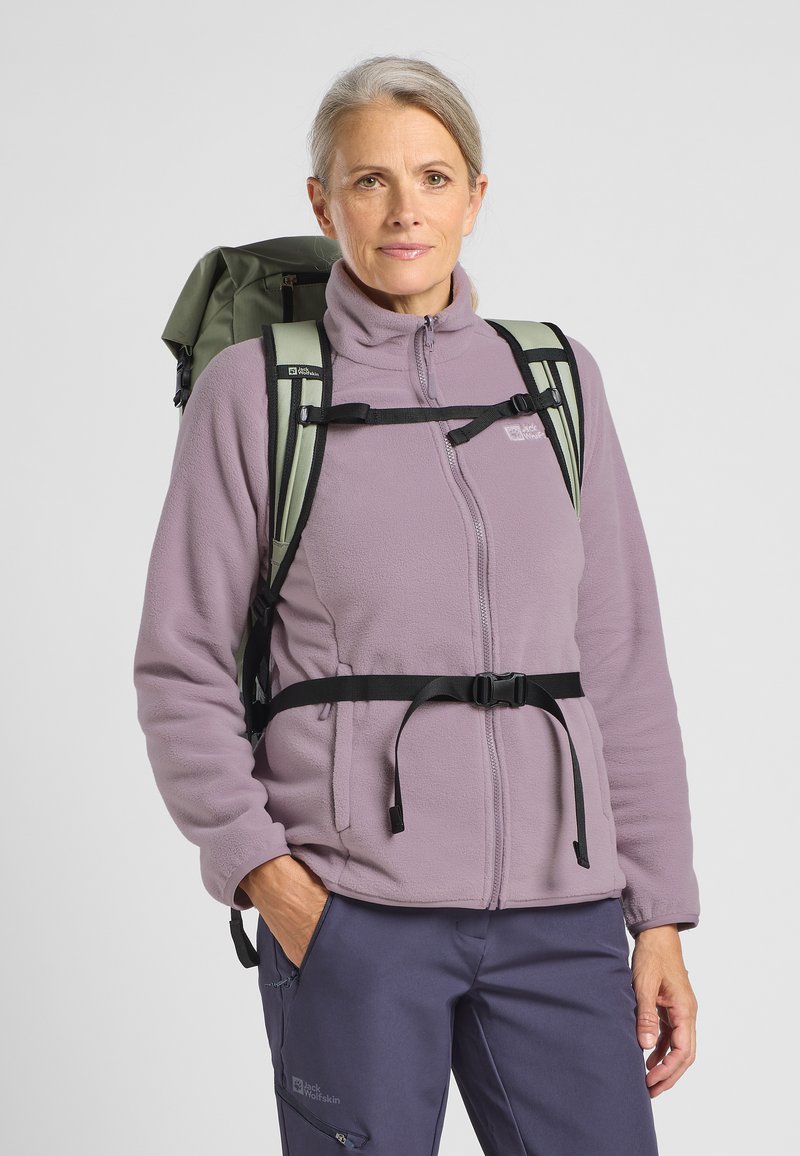 Donna matura che indossa una giacca in pile viola e pantaloni blu con uno zaino verde, in piedi con una mano in tasca davanti a uno sfondo grigio.