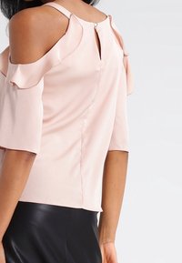 Blusa rosa pallido con spalle scoperte, dettagli a volant e maniche corte. Presenta un'apertura a chiave sul retro con chiusura a bottone. Texture liscia.